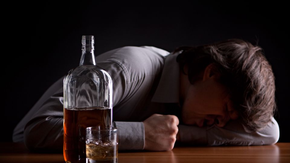 Egy férfi az asztalon fekszik, mellette üveg alkohol és pohár.