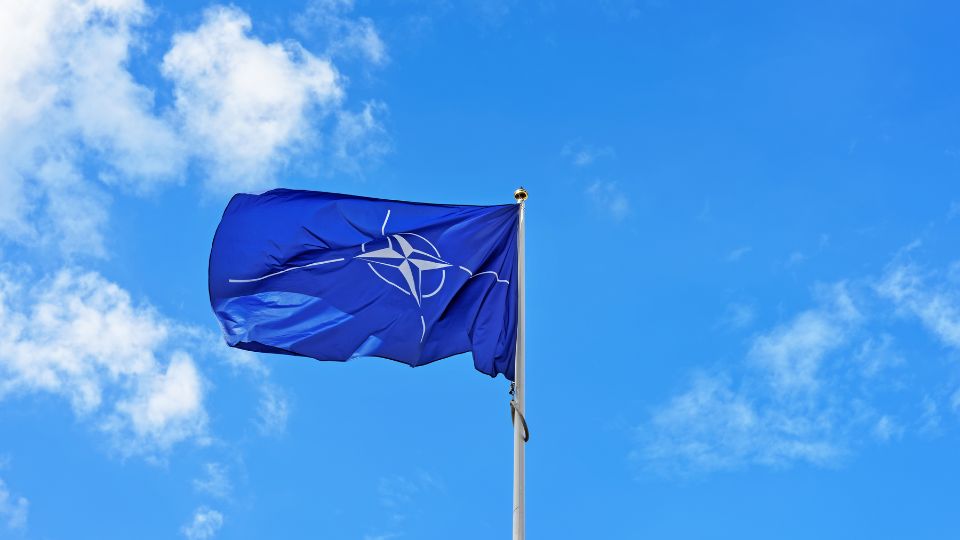 A NATO zászlaja lobog a kék égen, szimbolizálva a szövetség erejét.