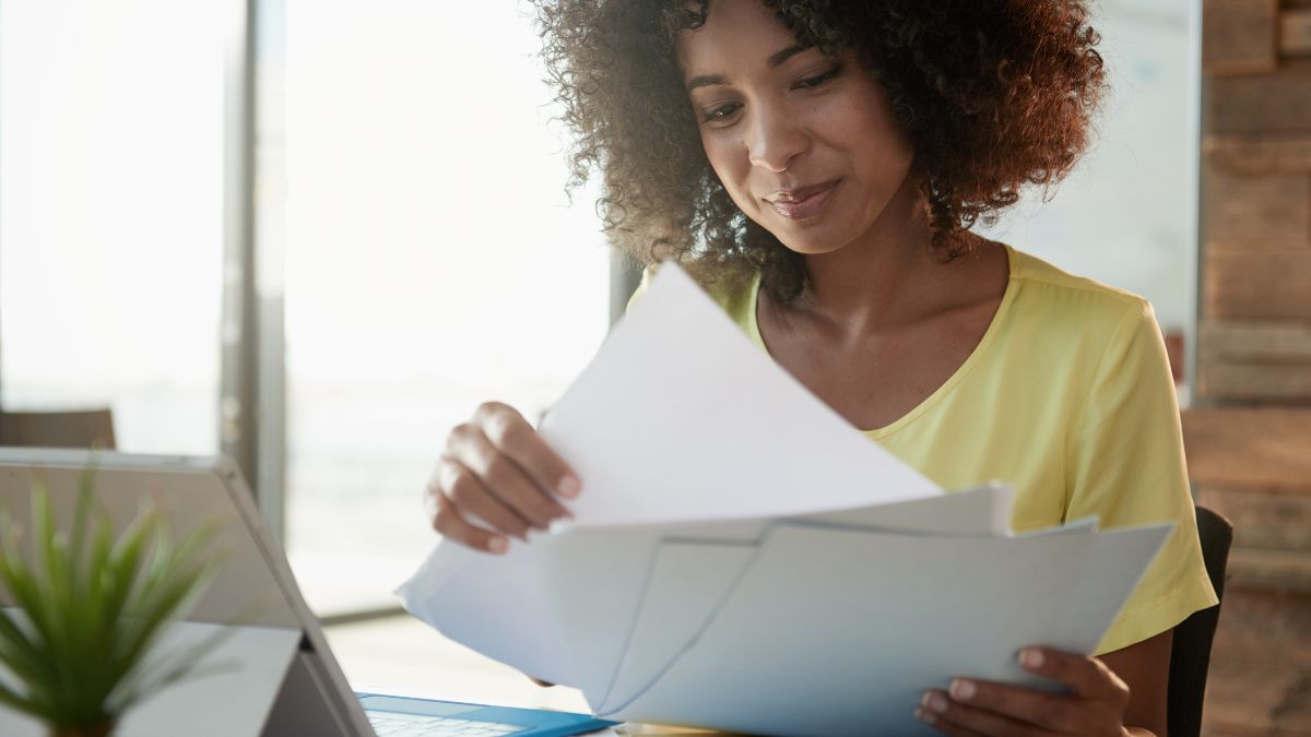 Egy fiatal nő iratokat nézeget, miközben egy laptop előtt ül.