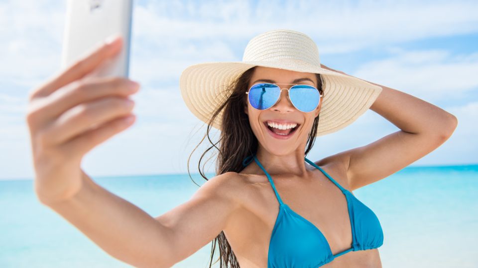 Egy fiatal nő napernyő alatt, bikiniben, mosolyogva készít szelfit a tengerparton.